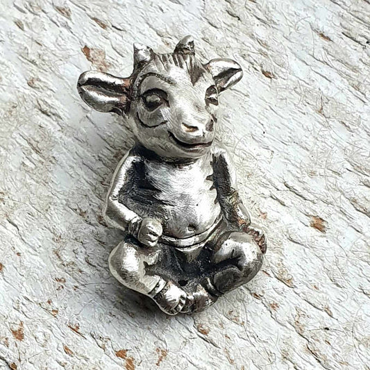 A silver-colored metal necklace in the shape of a stylized goat with human-like features, set against a wooden background.
