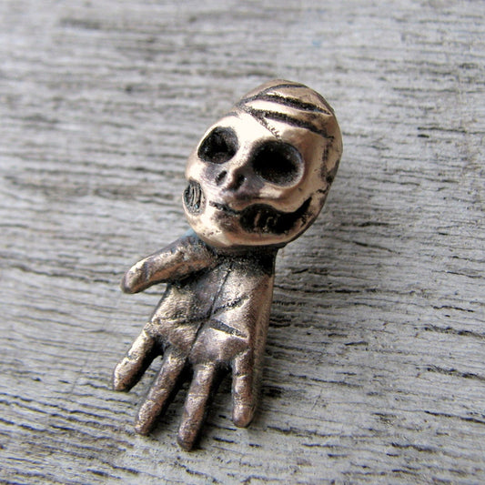 A bronze sculpture of a skeleton hand with a skull head, designed as a charm or pendant.