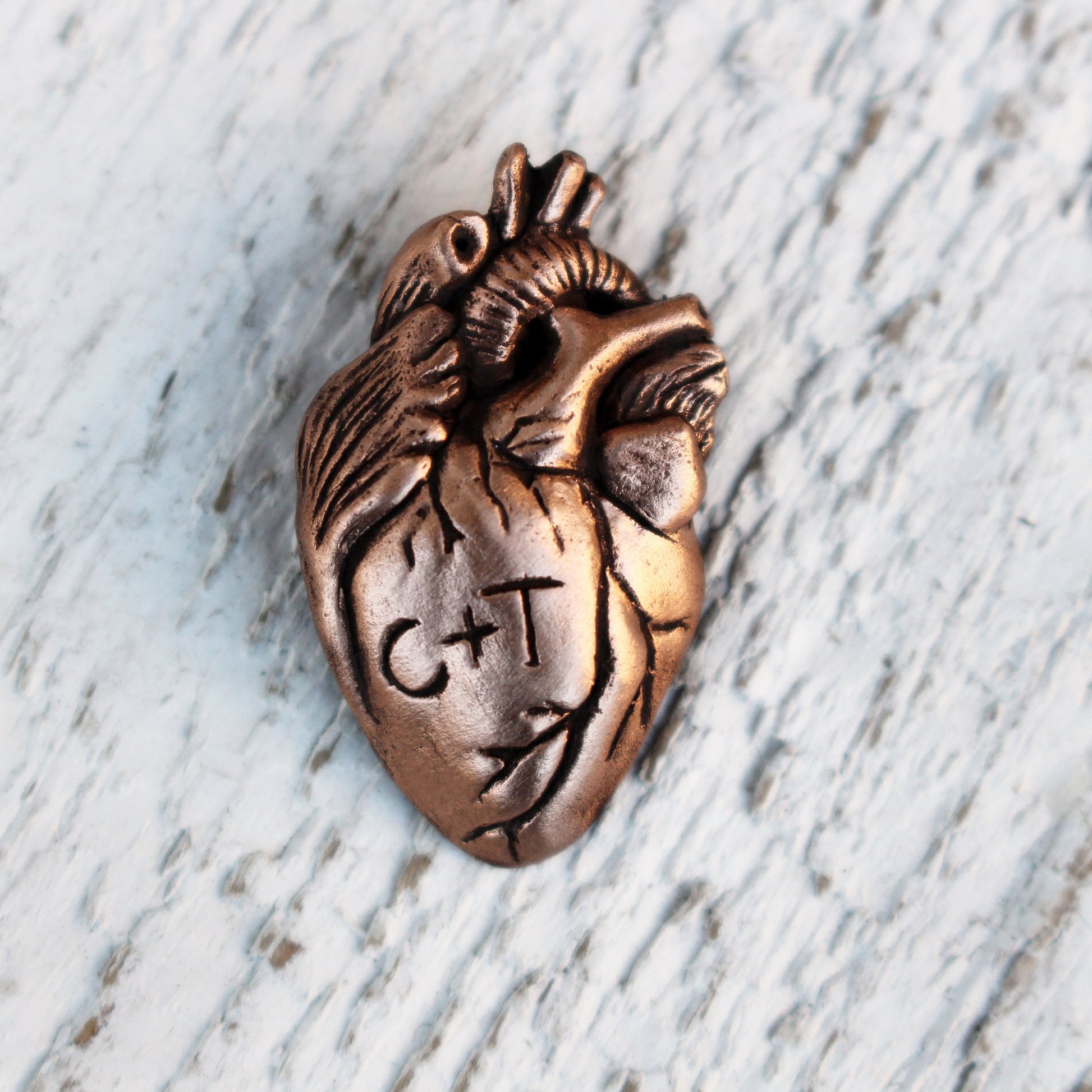 A custom anatomical heart necklace with the initials 'C+T' engraved on it, displayed against a wooden background.