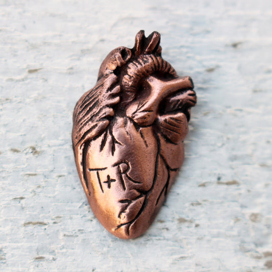 A bronze-colored anatomical heart brooch with personalized initials.