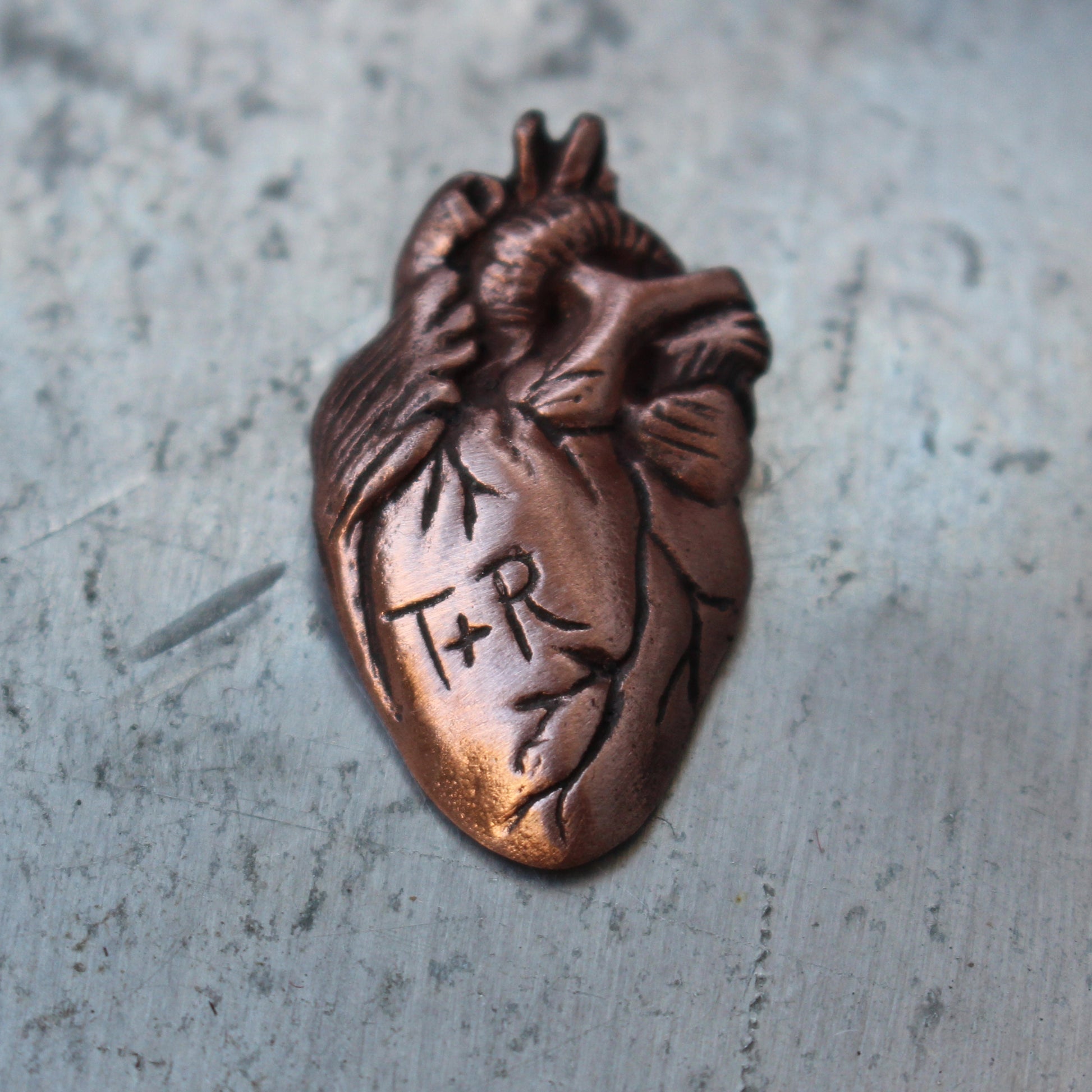 A custom copper brooch in the shape of an anatomical heart with engraved initials 'T&R' on the front.