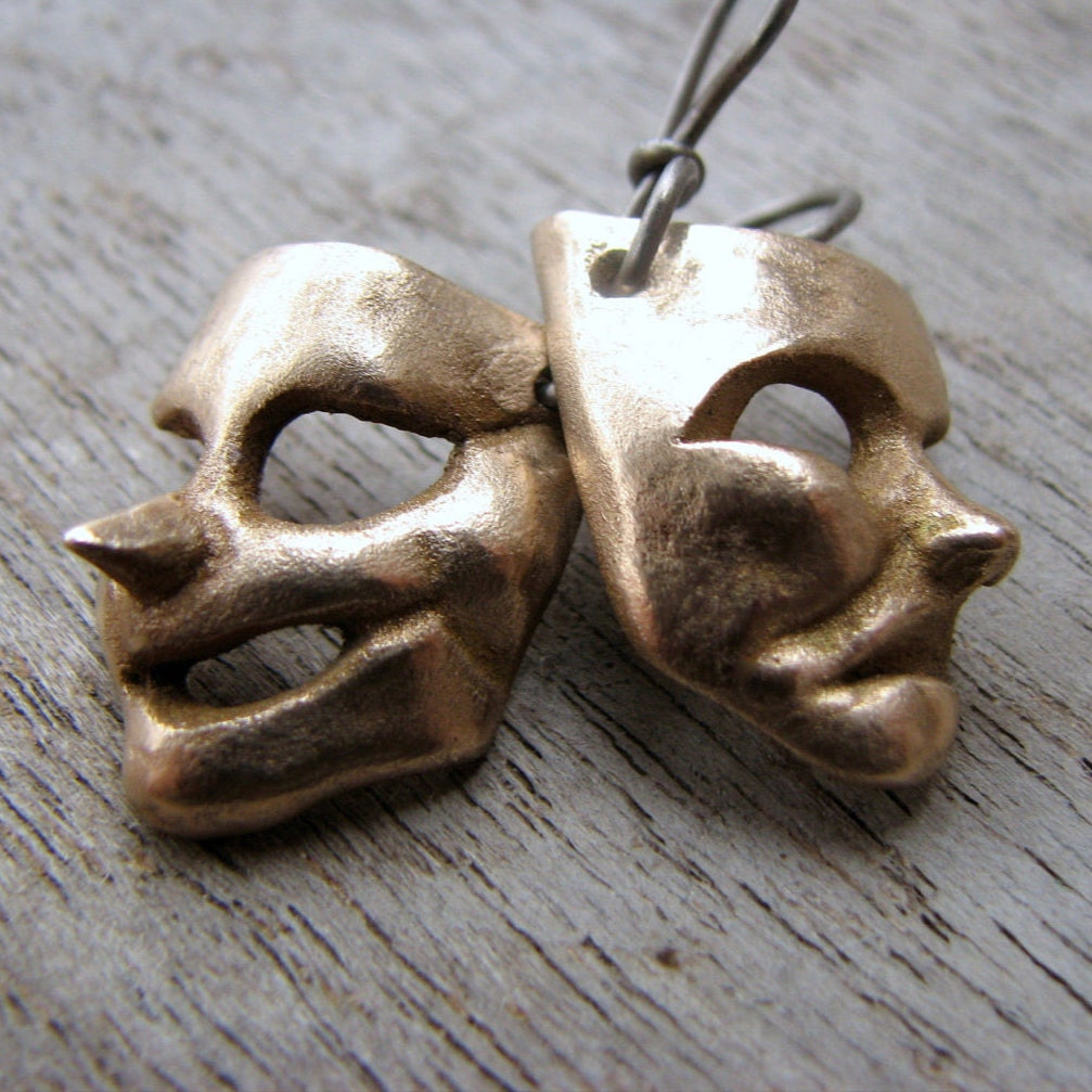 A pair of bronze theater mask earrings, one depicting a comedy mask and the other a tragedy mask, placed on a wooden surface.