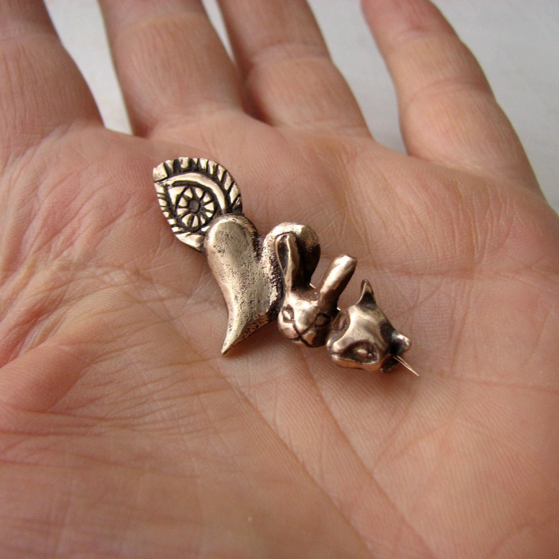 A bronze-colored brooch with a design that includes a rabbit and a cat, held in a person's hand.