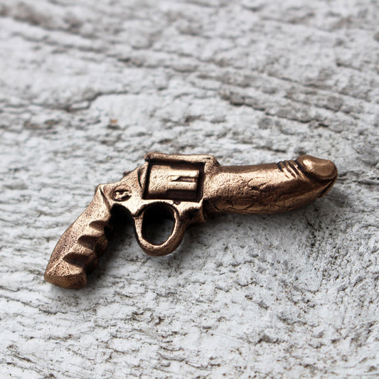 A bronze-colored miniature revolver-shaped pendant with phallic features, displayed on a textured background.