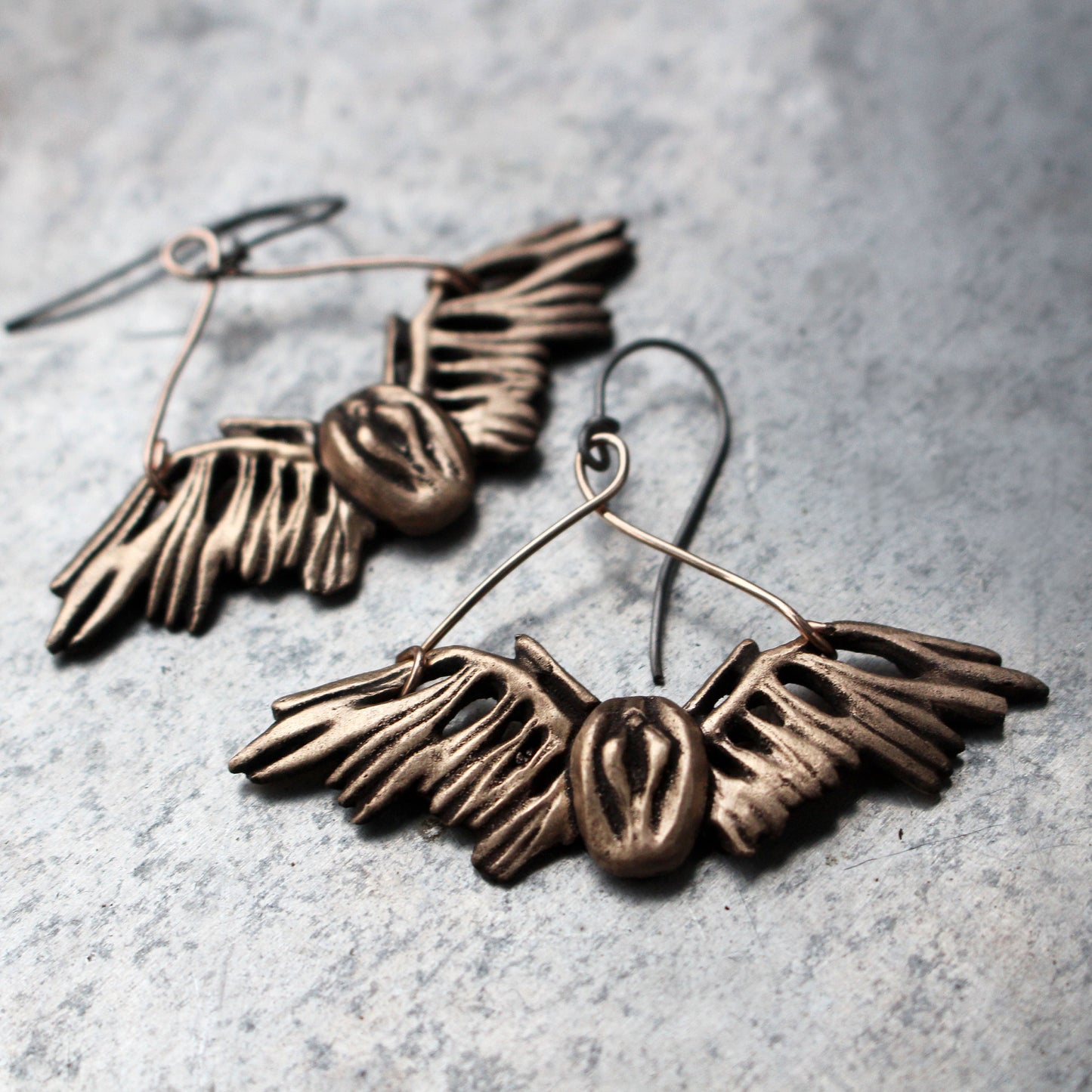 This image displays a pair of distinctive earrings featuring winged, sculpted designs that resemble female genitalia. Crafted from bronze, these intricate earrings dangle elegantly from niobium wire hooks, which are ideal for individuals with metal allergies.