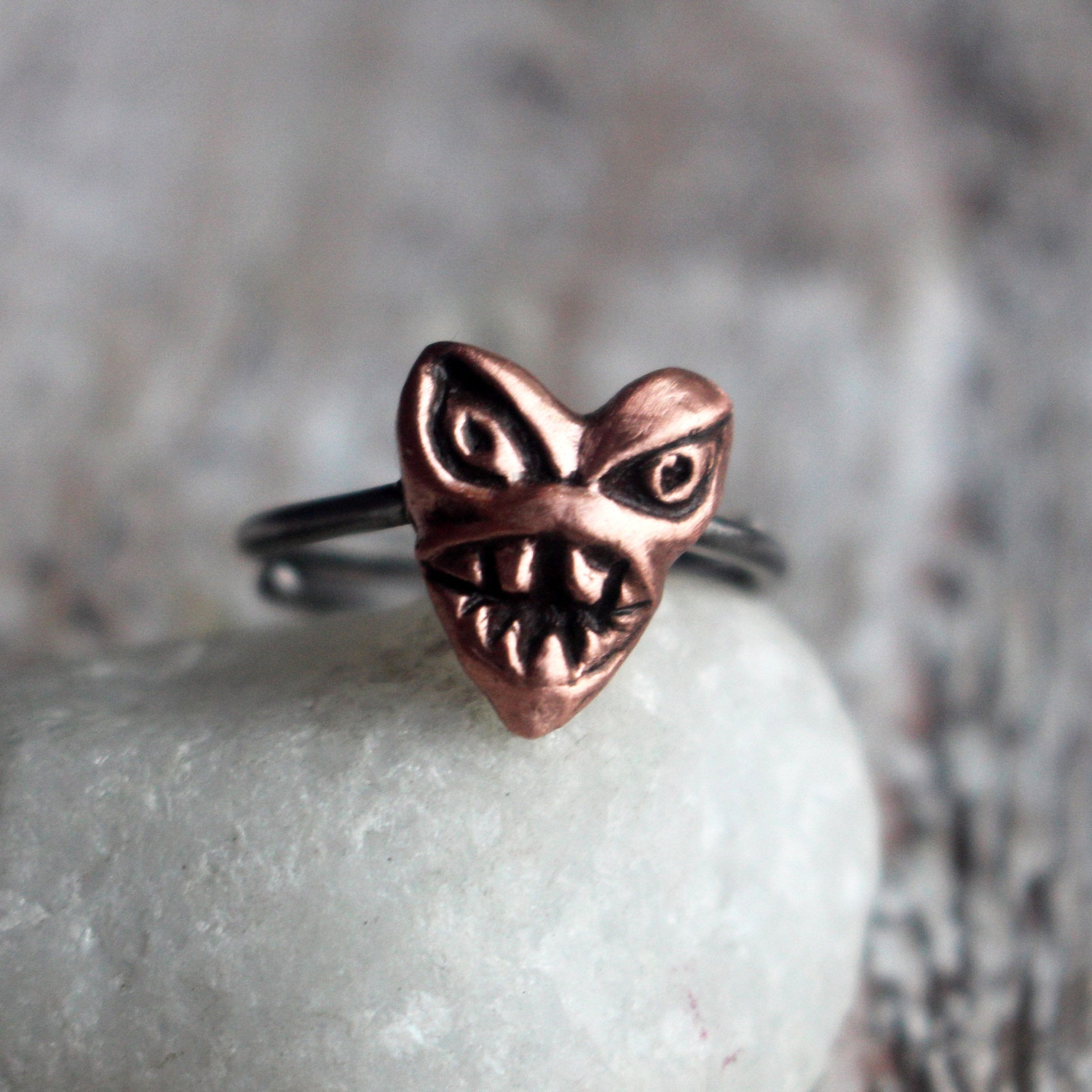 A ring with a pink heart-shaped copper monster sculpted on top, displayed on a white stone with a wooden background.