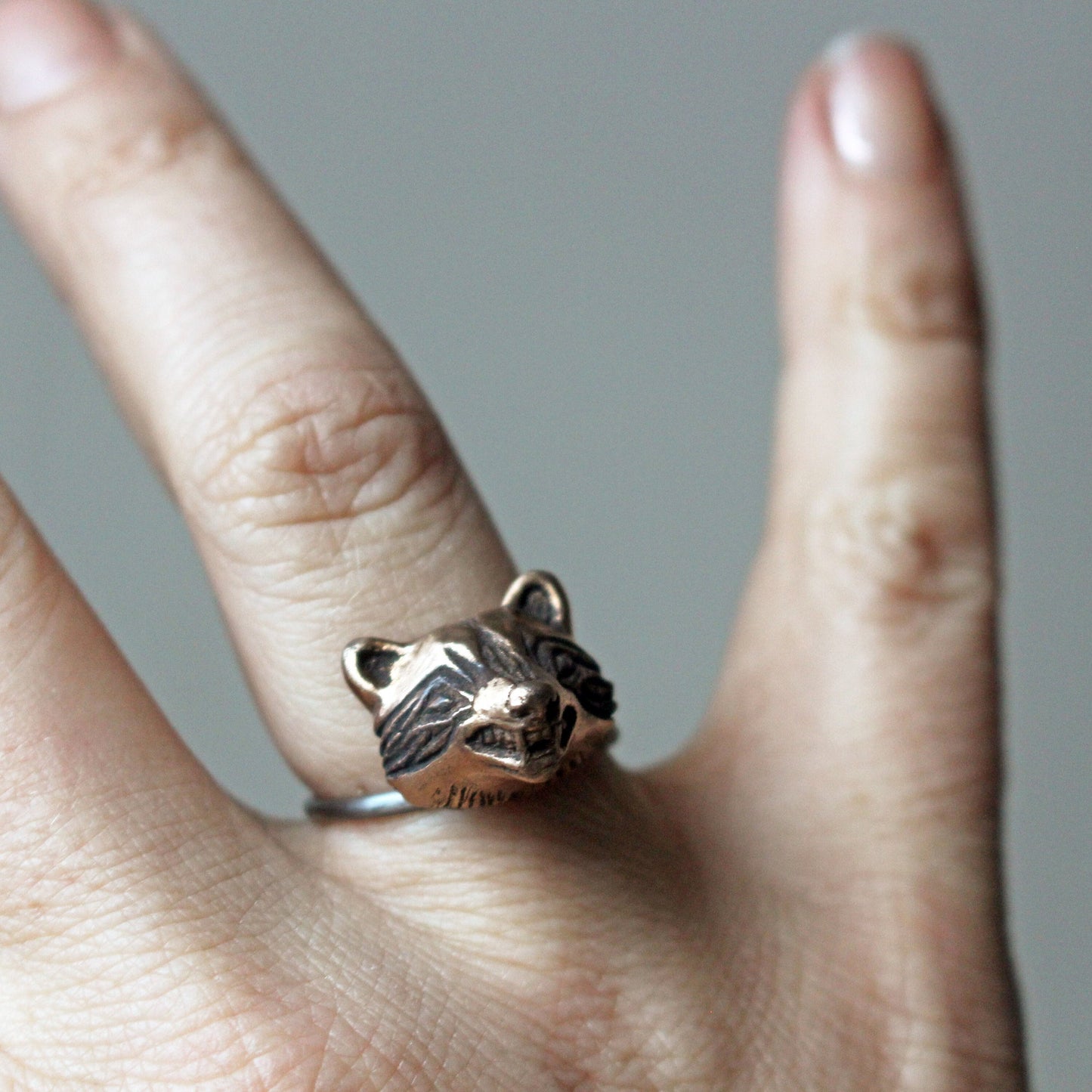 A bronze-colored raccoon head ring on a person's finger, showcasing the ring's detail and the animal's facial features.
