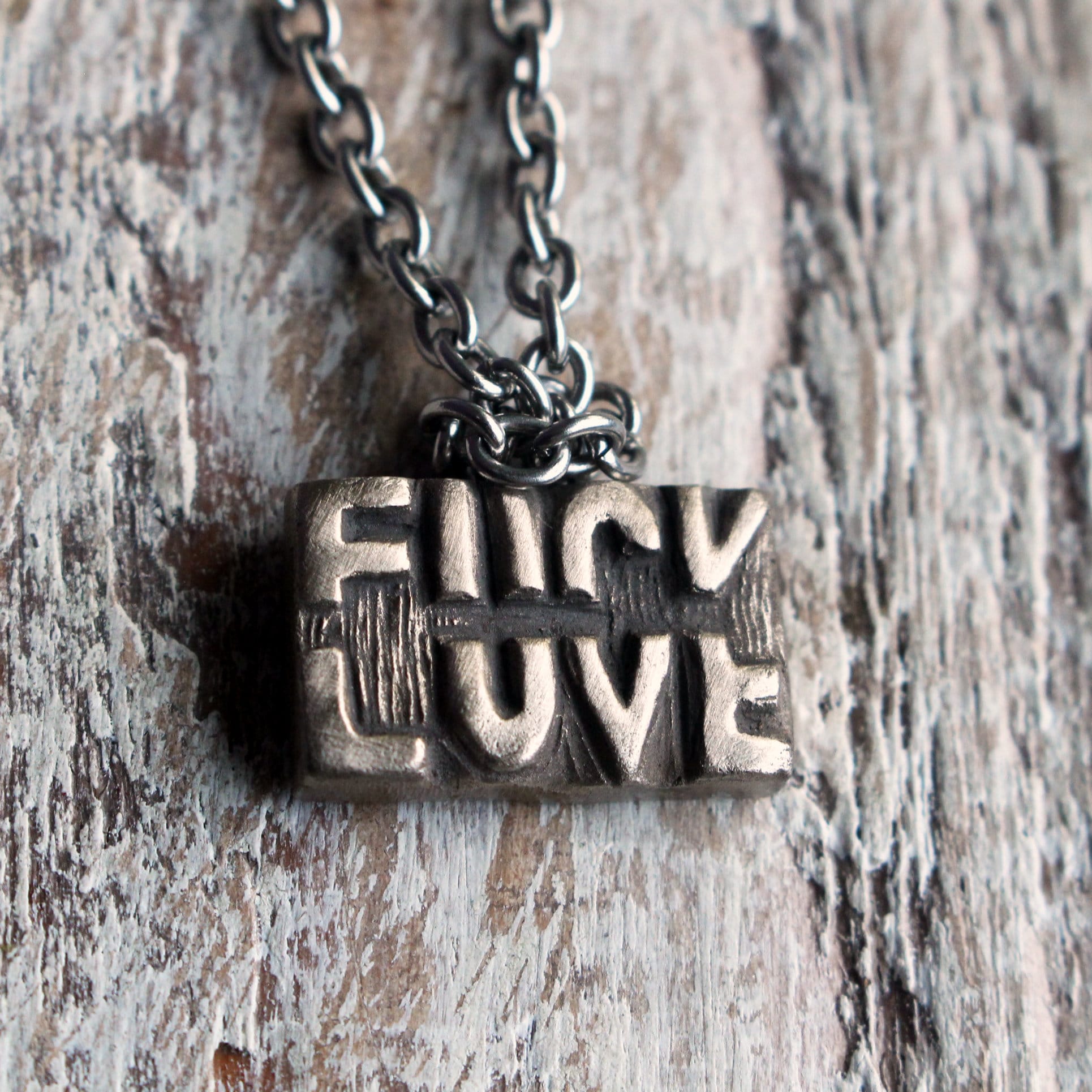 A bronze pendant necklace with the phrase 'FUCK LOVE' sculpted on the pendant, attached to a stainless steel chain, displayed against a wooden background.
