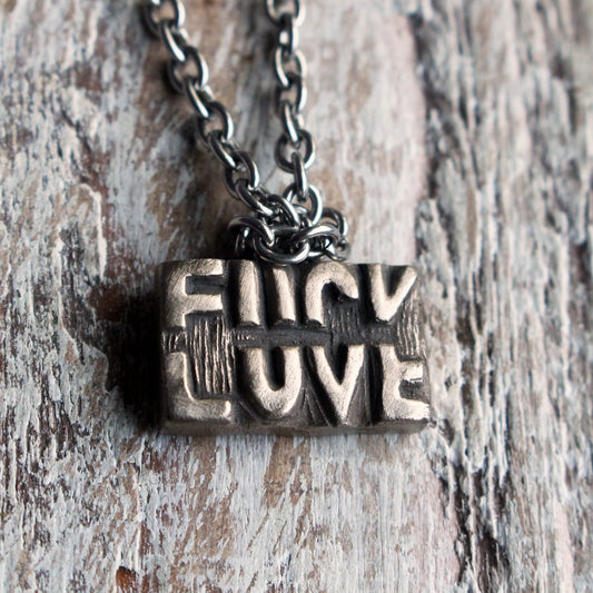 A bronze pendant necklace with the phrase 'FUCK LOVE' sculpted on the pendant, attached to a stainless steel chain, displayed against a wooden background.