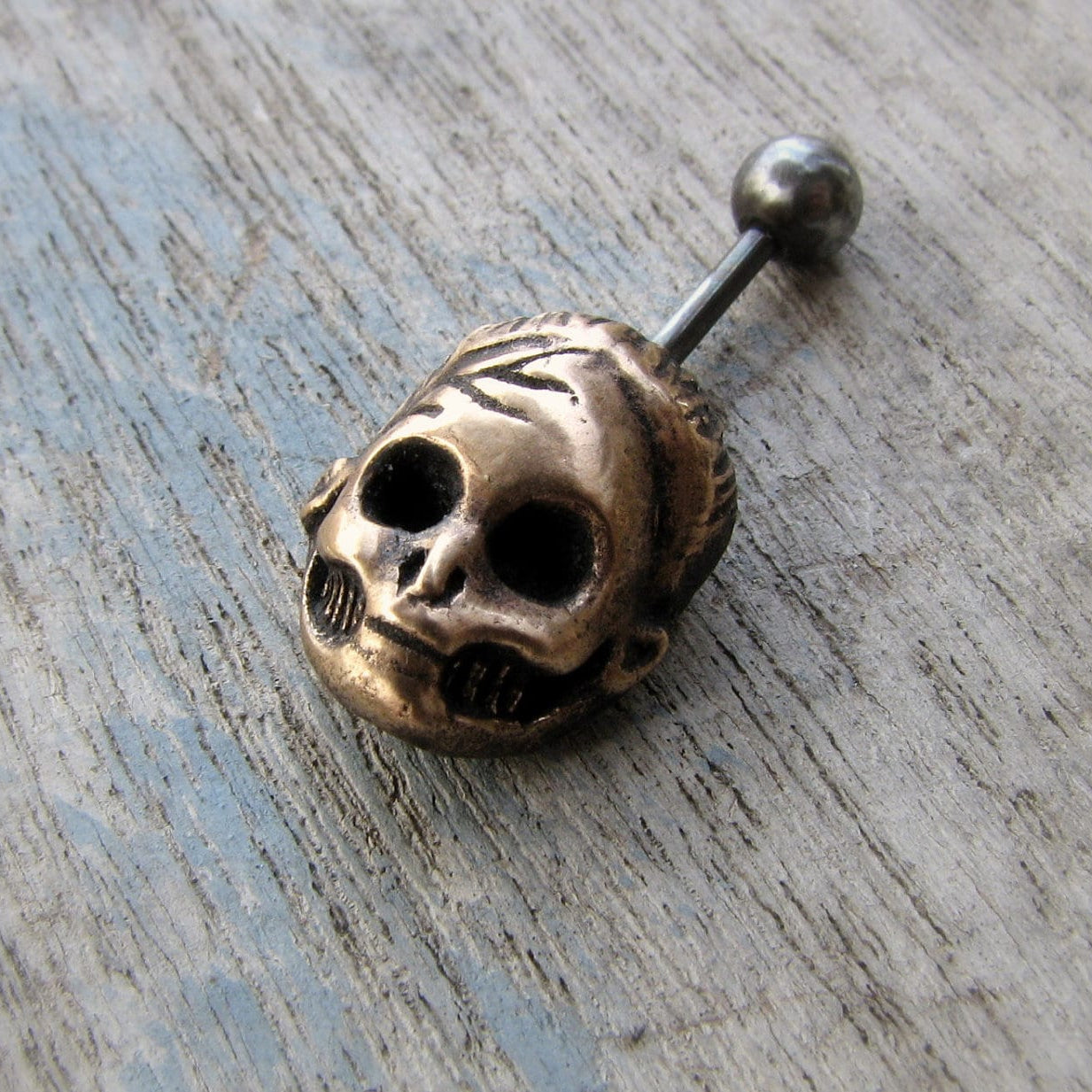 A bronze belly button ring with a skull design, made of surgical steel, displayed on a wooden surface.