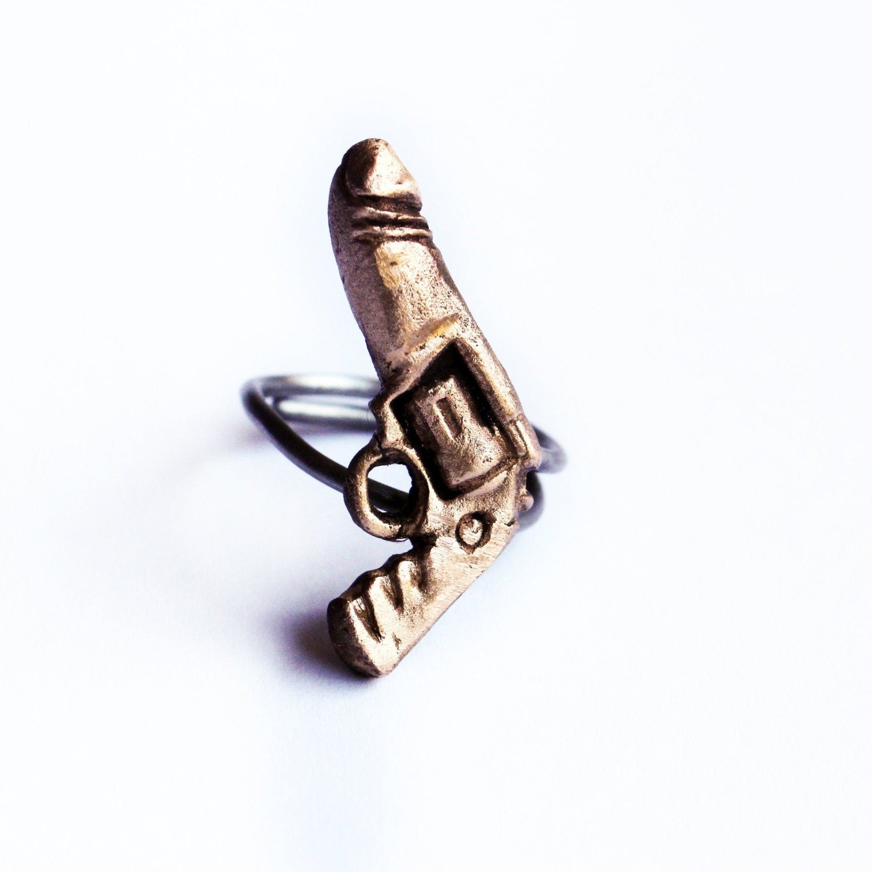 A bronze ring with a sculpted design resembling a penis gun, set on a stainless steel band. The ring is displayed against a white background.
