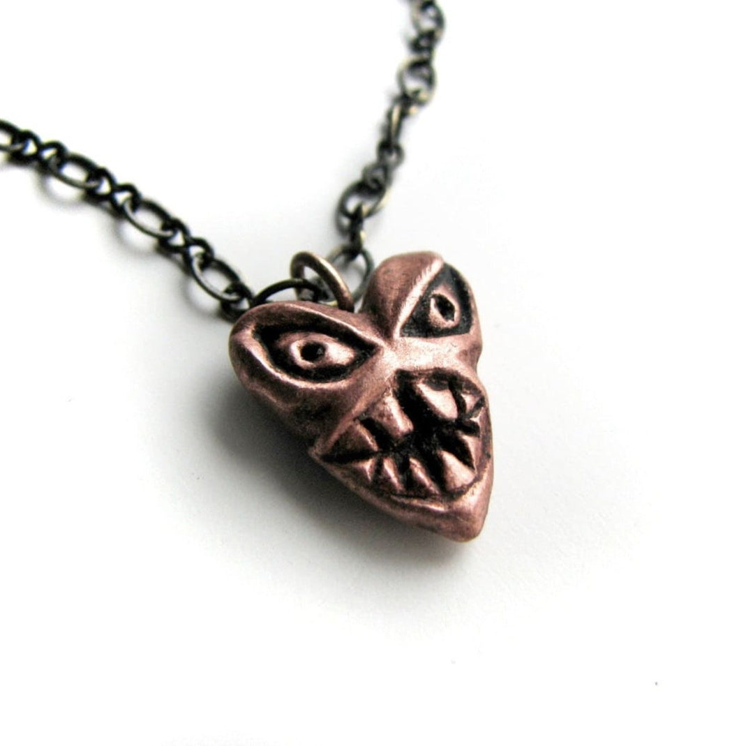 A copper pendant in the shape of a heart with a monster face design, attached to a black chain.