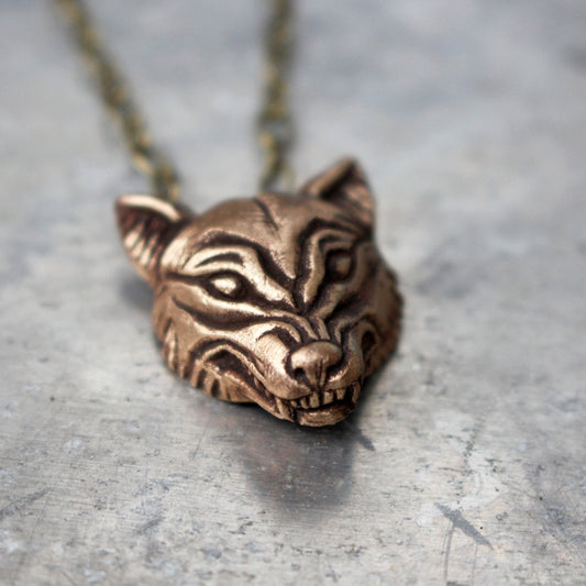 Bronze wolf head pendant necklace on a chain, displayed against an oxidized metal background.