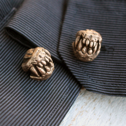 A pair of bronze cuff links designed to resemble animal or monster skulls, placed on a striped fabric surface.
