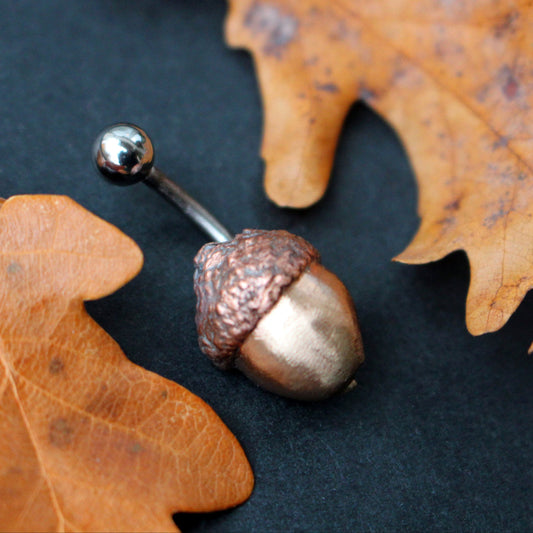 Acorn belly button ring with a bronze nut, copper cap, and a curved barbell, displayed on a dark surface with autumn leaves.