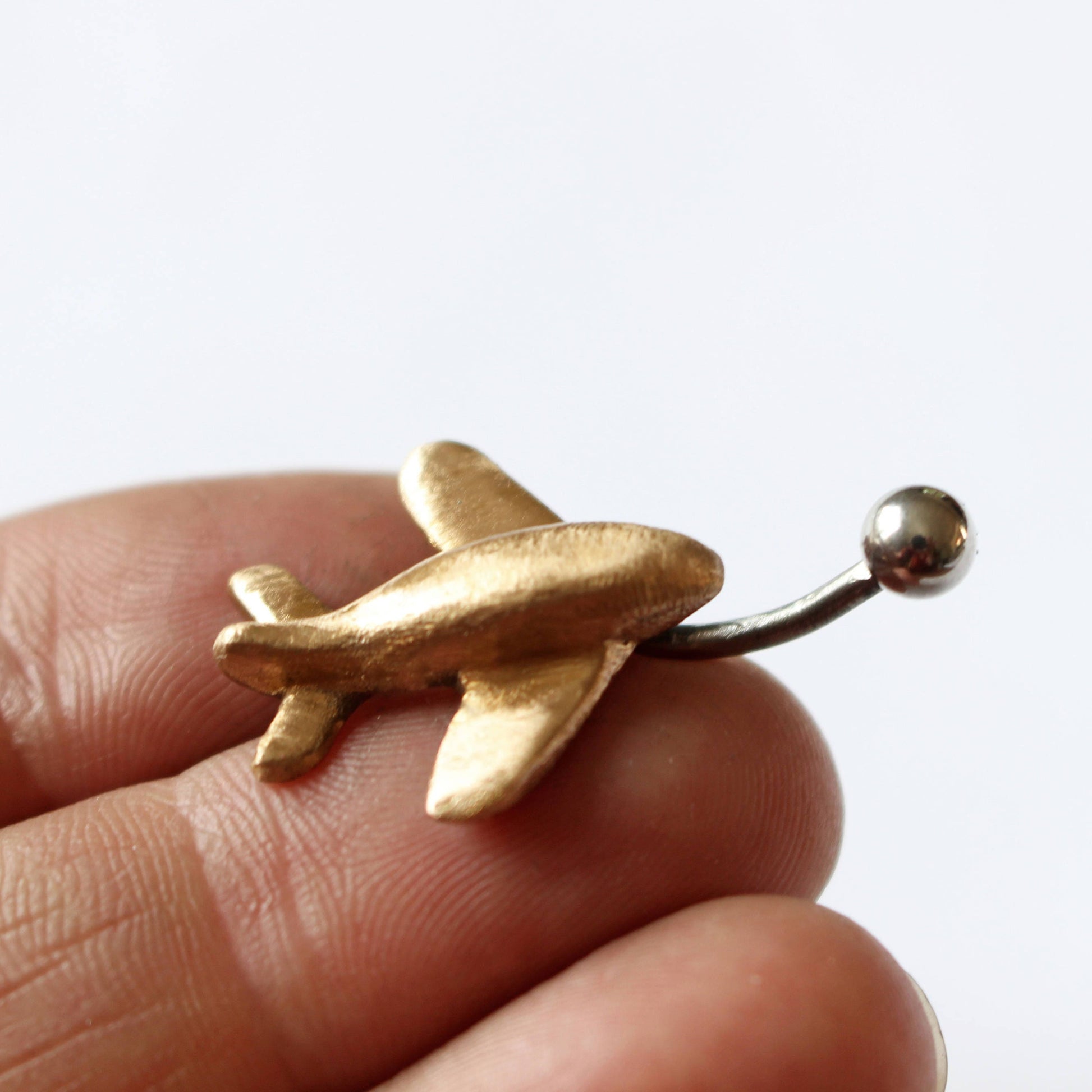 A bronze-colored airplane-shaped belly button ring held between fingers, with a metallic barbell and a small ball attachment.