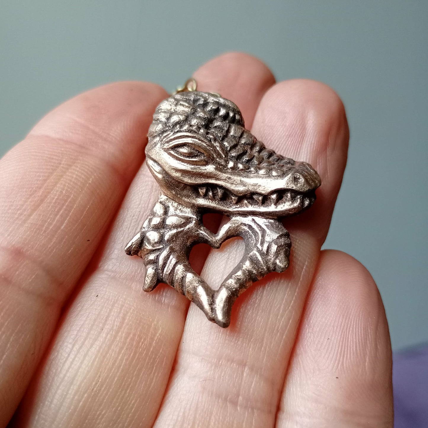 Bronze alligator/crocodile pendant held between fingers against a neutral background, the crocodilia makes heart hands