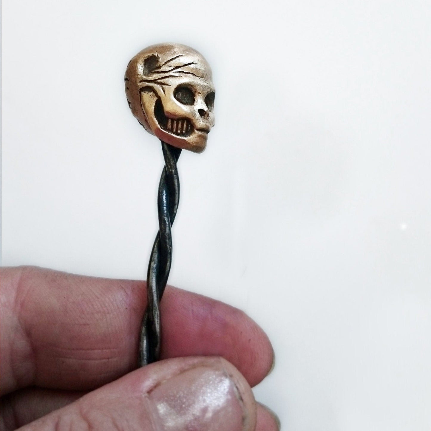 A bronze hair stick with a skull design, held between fingers against a white background.