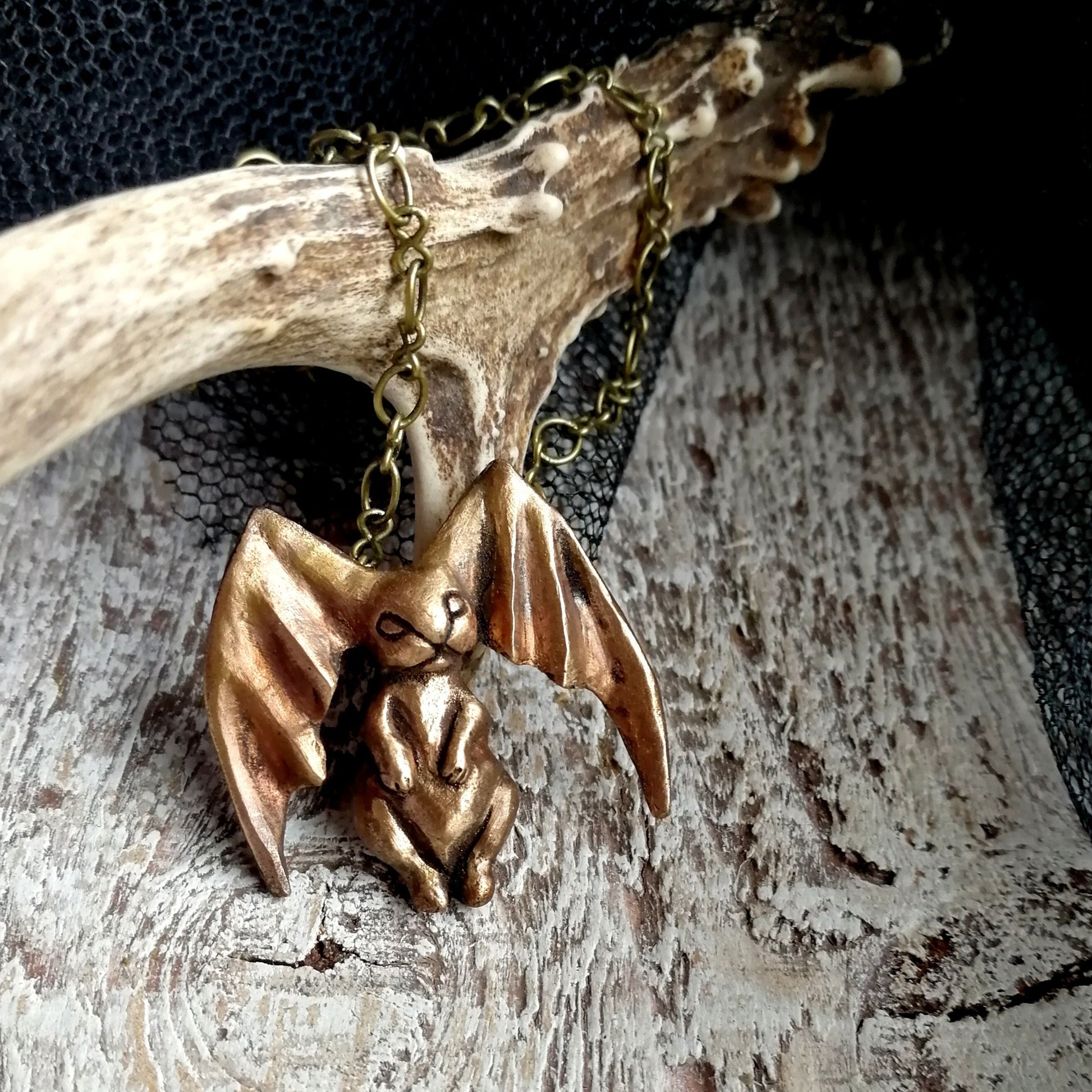 A bronze pendant sculpture of a bunny with bat wings as ears, displayed on a chain against 