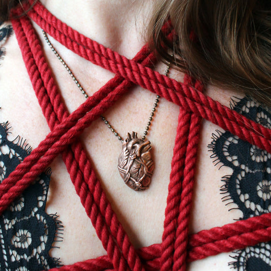 A realistic heart-shaped pendant necklace made from copper clay, bound with red rope, displayed on a person's chest.