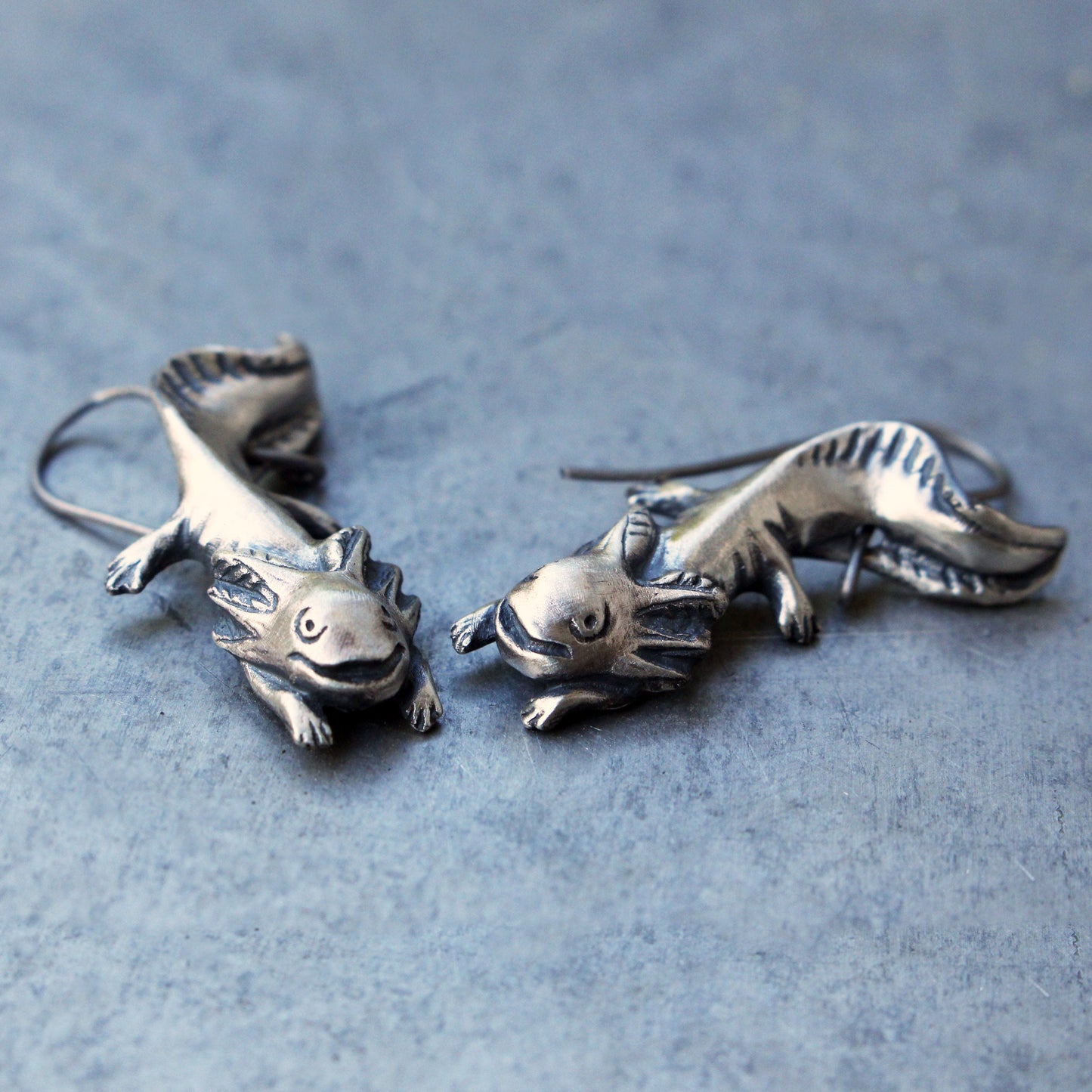 A pair of handmade axolotl, mudpuppy earrings made from bronze and sterling silver, displayed on a textured background.