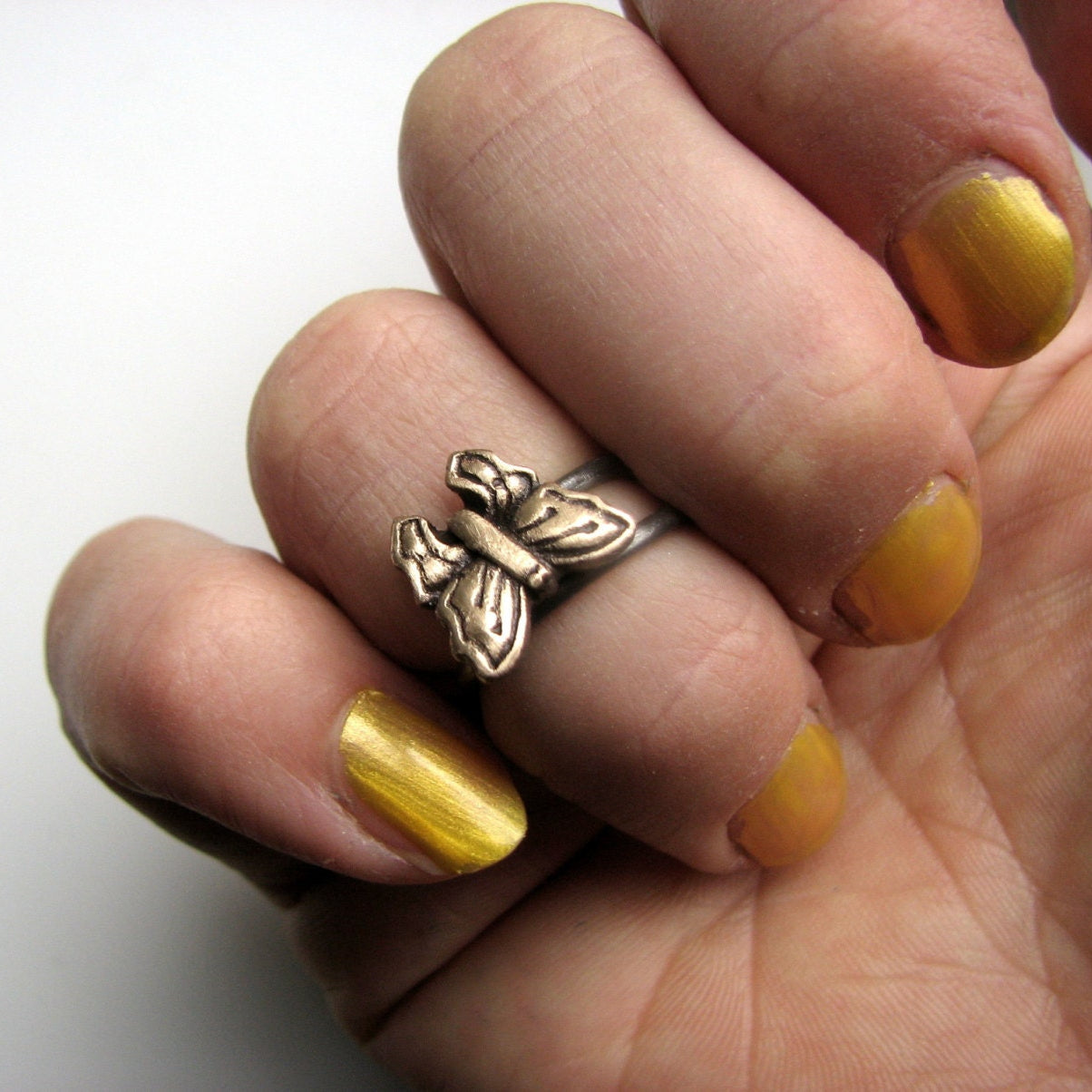 Hand wearing a bronze butterfly ring with yellow nail polish on a white background