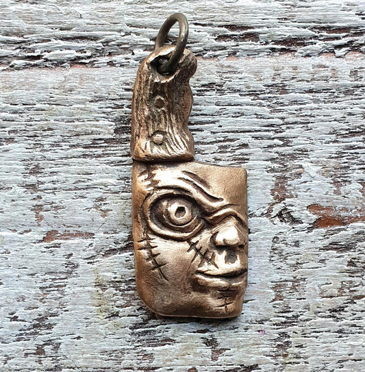 Bronze-colored pendant necklace with a sculpted face resembling the horror doll Chucky on the blade, displayed against a wooden background.