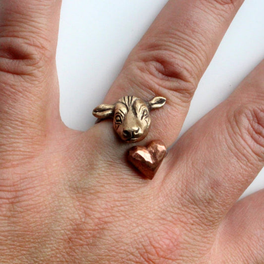 An adjustable ring with a bronze cow head on one end and a pink copper heart on the other, displayed on a person's hand.