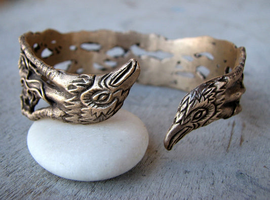 A bronze cuff bracelet featuring two crow or raven bird heads with delicate filigree work displayed on a white stone.