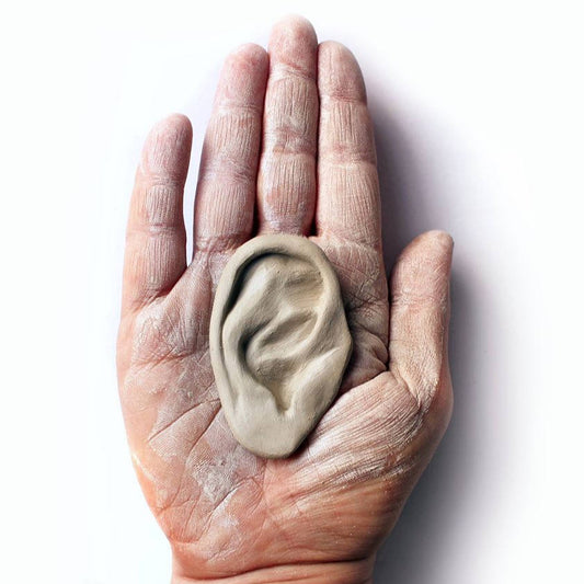 A ceramic sculpture of an ear displayed on a hand, with a matte white finish.