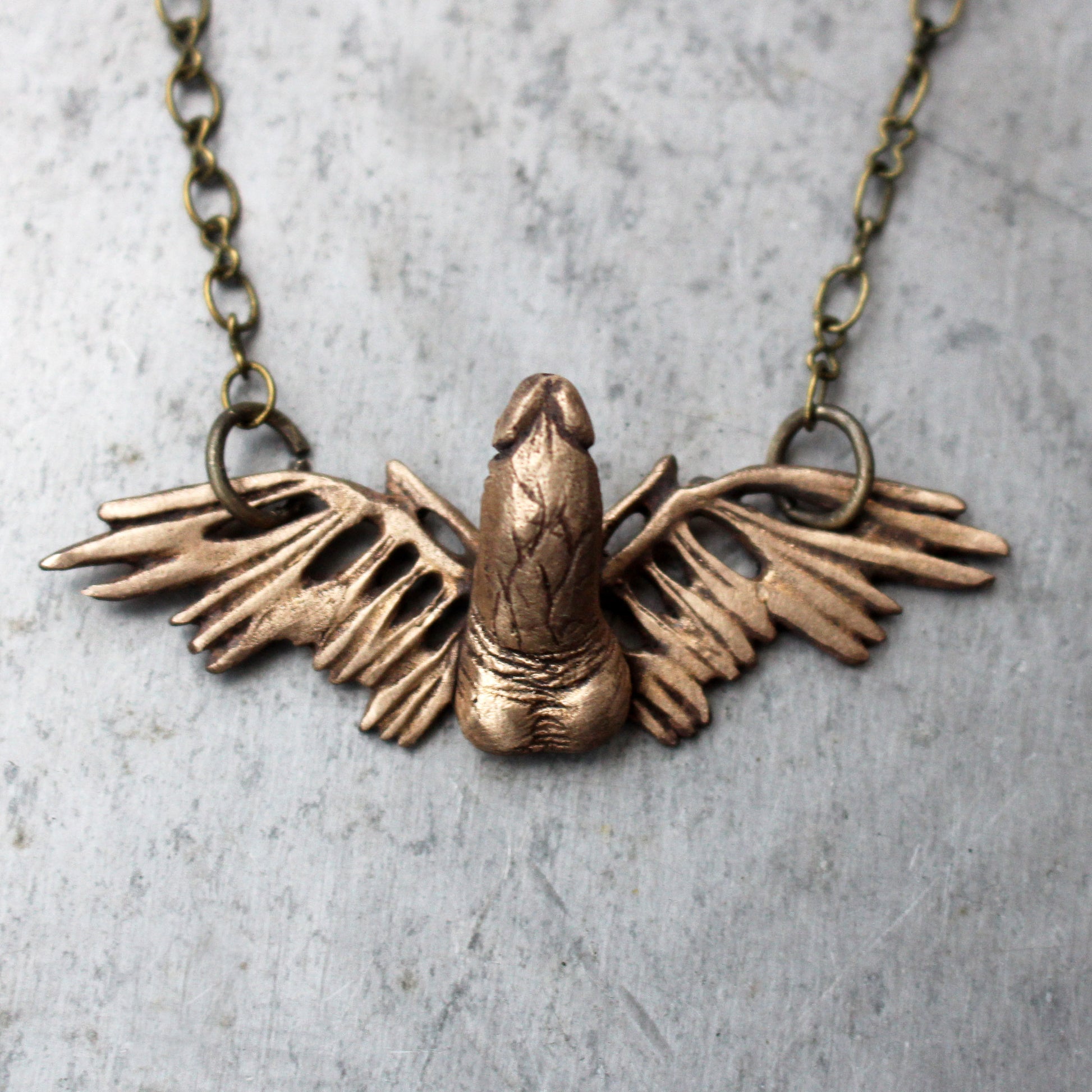 A bronze winged phallus pendant necklace with adjustable brass chain displayed against a textured background.