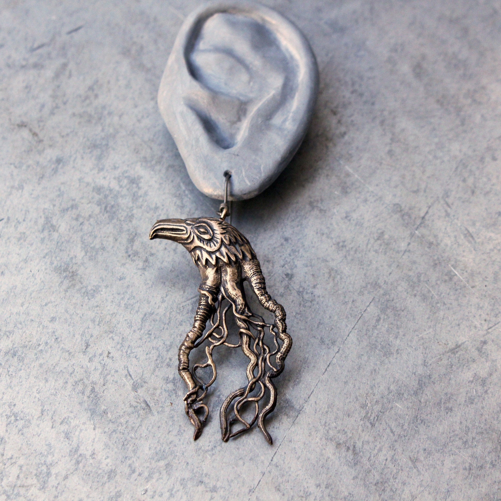 A bronze-colored raven-shaped earring with root or blood vein details, displayed against a grey background.