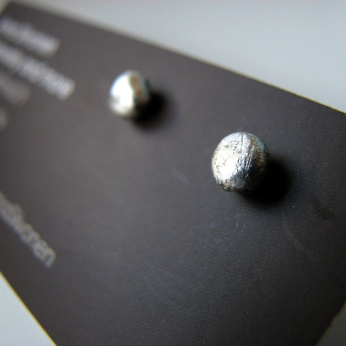 Close-up of a pair of silver pebble-shaped earrings with a scratched surface, displayed on a black card.