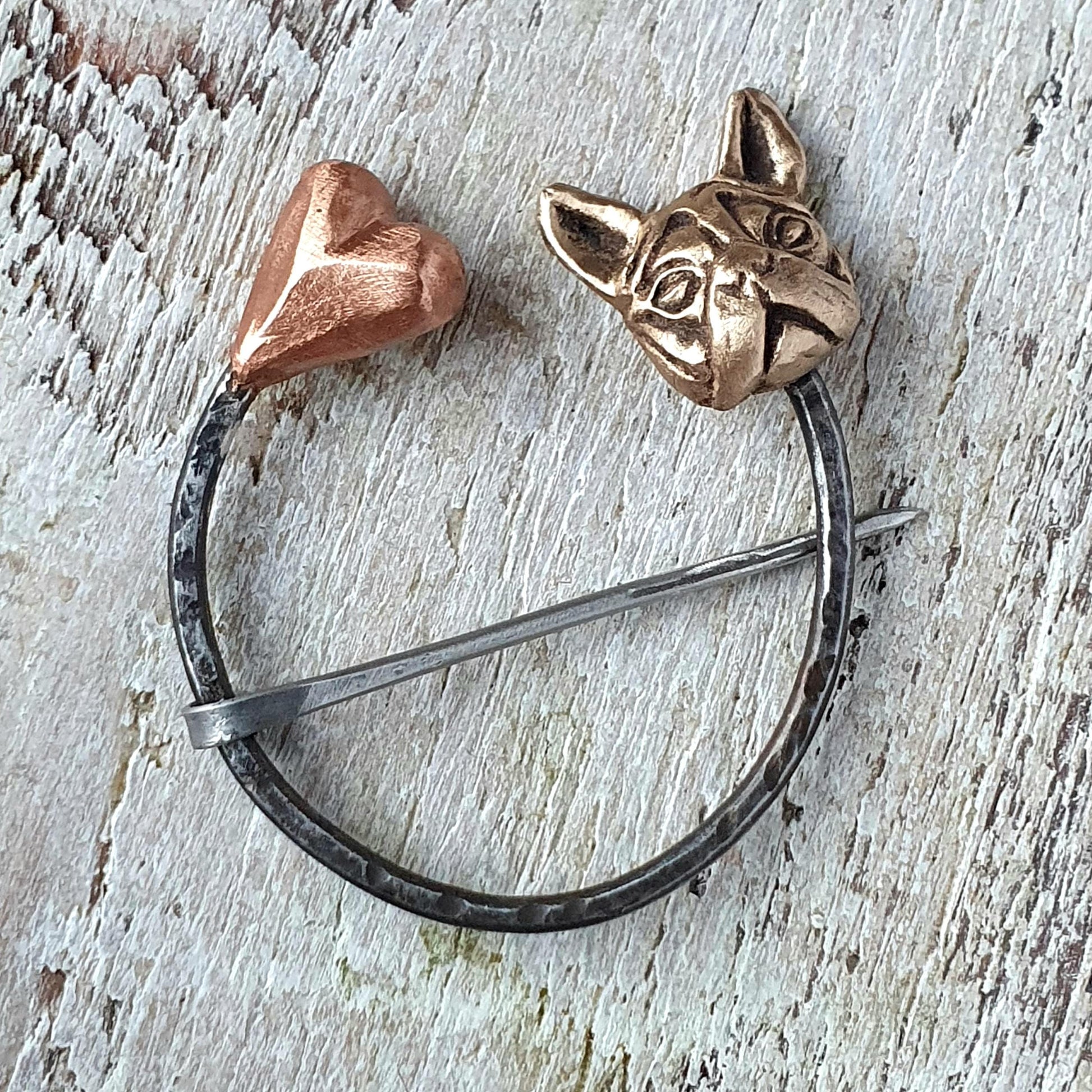 A handmade brooch featuring a bronze dog head and a pink copper heart, connected by a stainless steel circle and pin, set against a wooden background.
