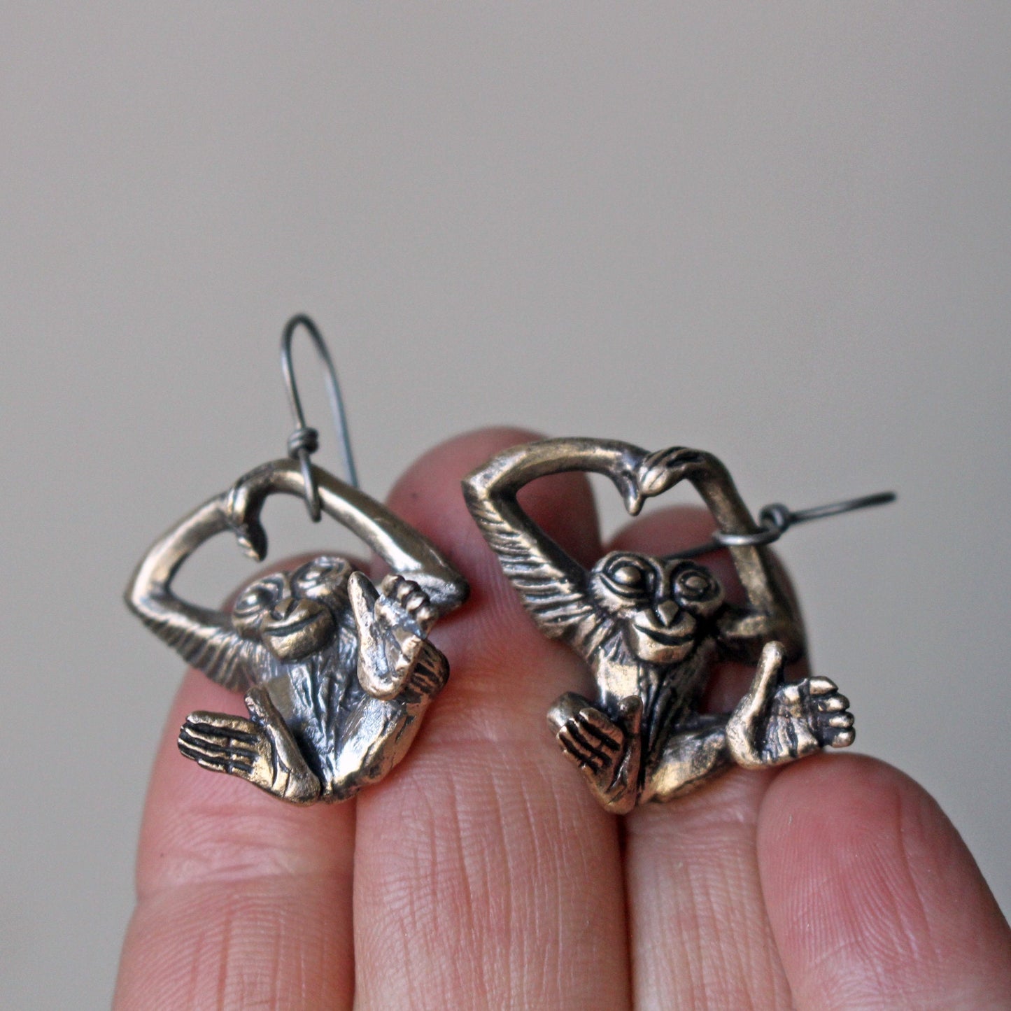 A pair of bronze-colored dangling earrings in the shape of monkeys with detailed feet and hands, displayed on a person's fingertip.