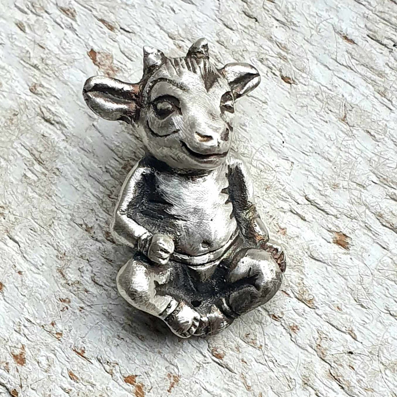 A silver-colored metal necklace in the shape of a stylized goat with human-like features, set against a wooden background.