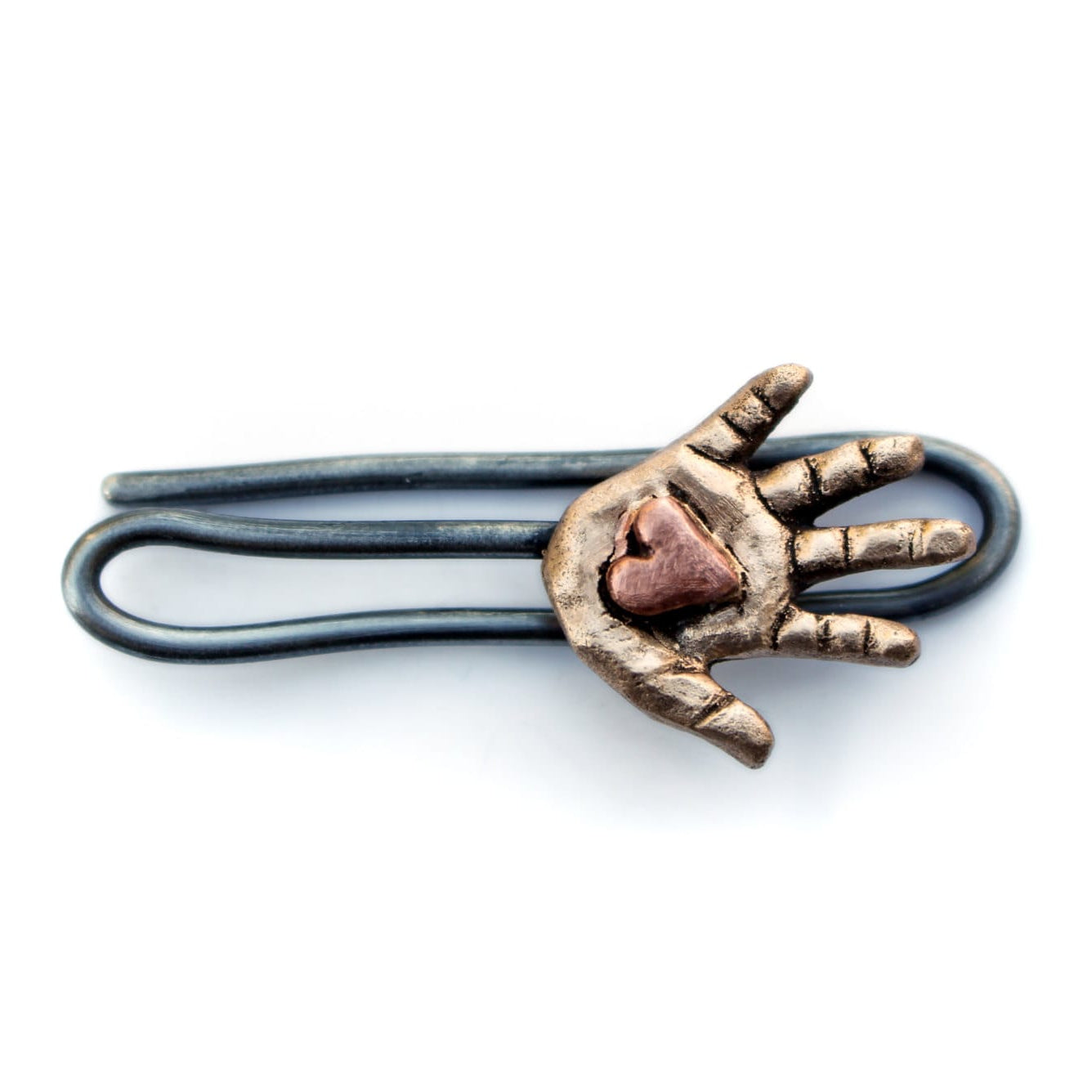 A tie clip with a bronze hand holding a pink copper heart in its palm, attached to a stainless steel wire pin.