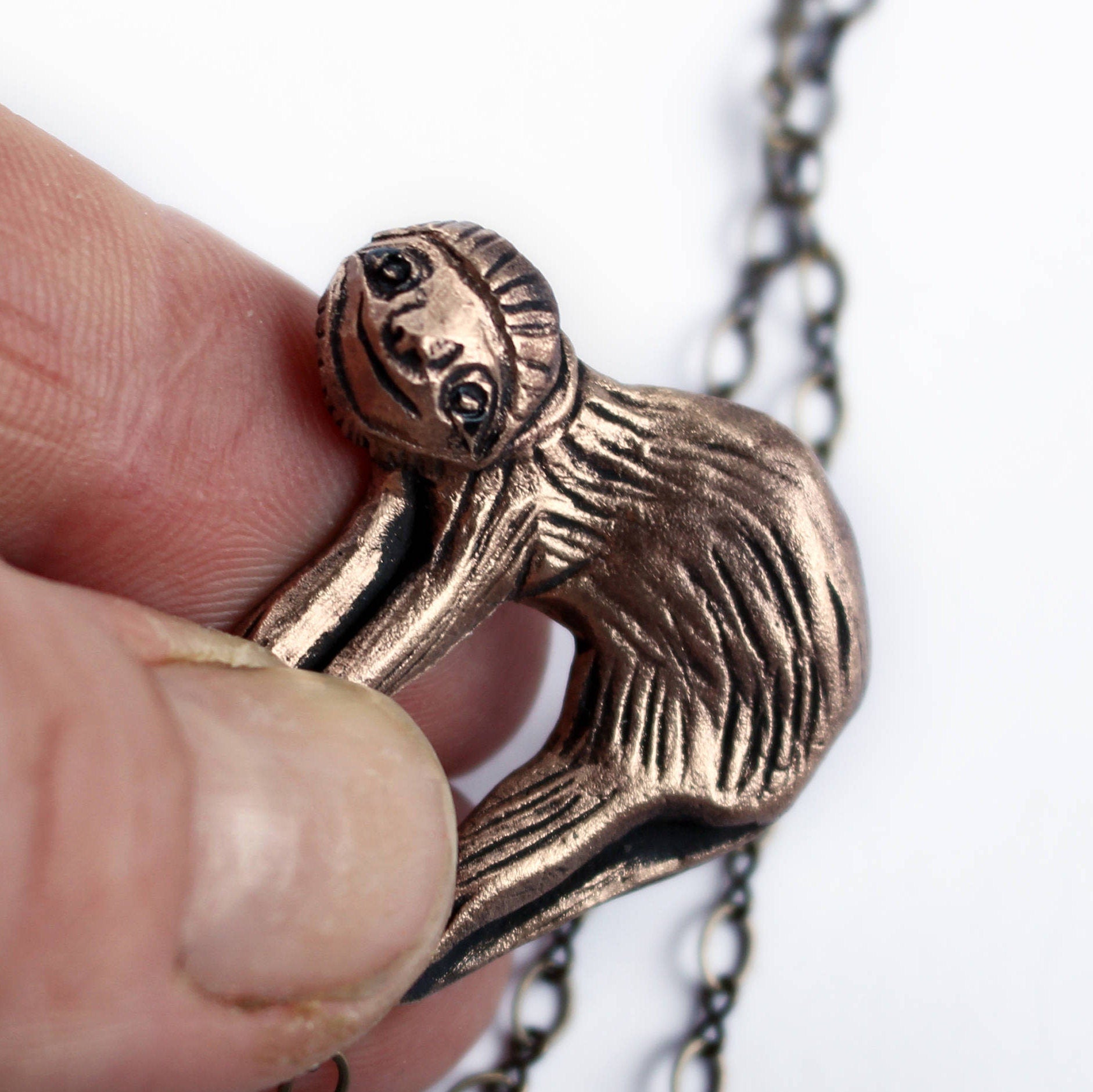 A bronze necklace featuring a smiling sloth sculpted as the pendant, held between fingers, with an adjustable brass chain in the background.