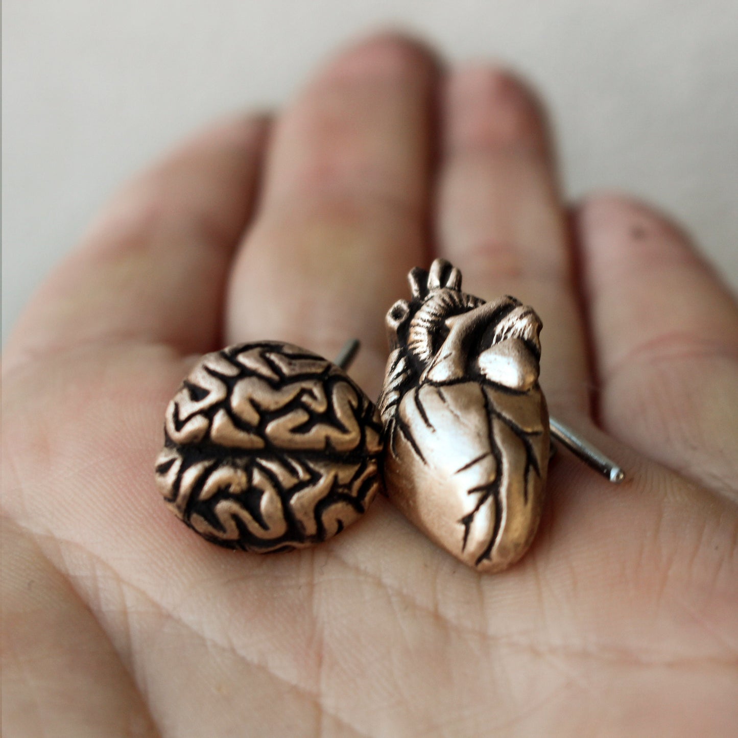 A pair of bronze cufflinks, one shaped like a heart and the other like a brain, held in a person's hand.