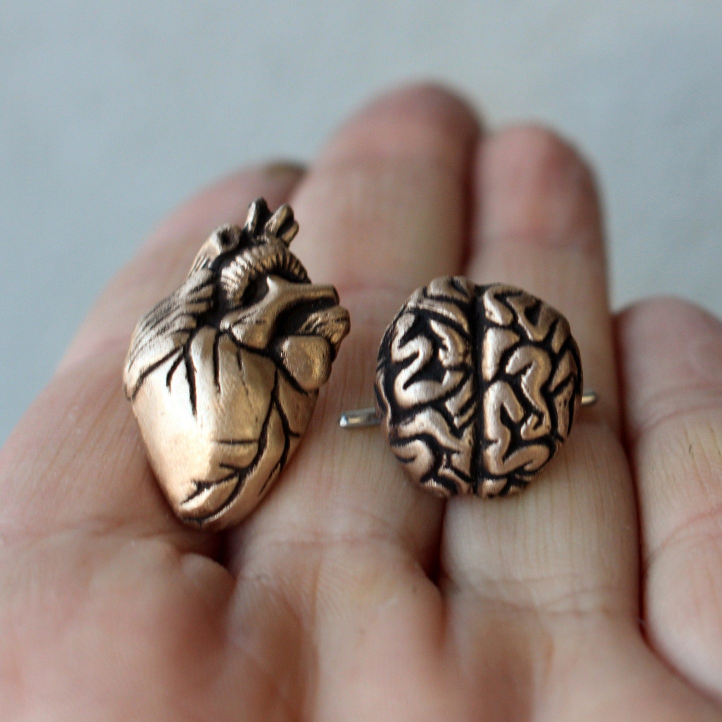 A pair of bronze cufflinks, one shaped like a heart and the other like a brain, held in a person's hand.