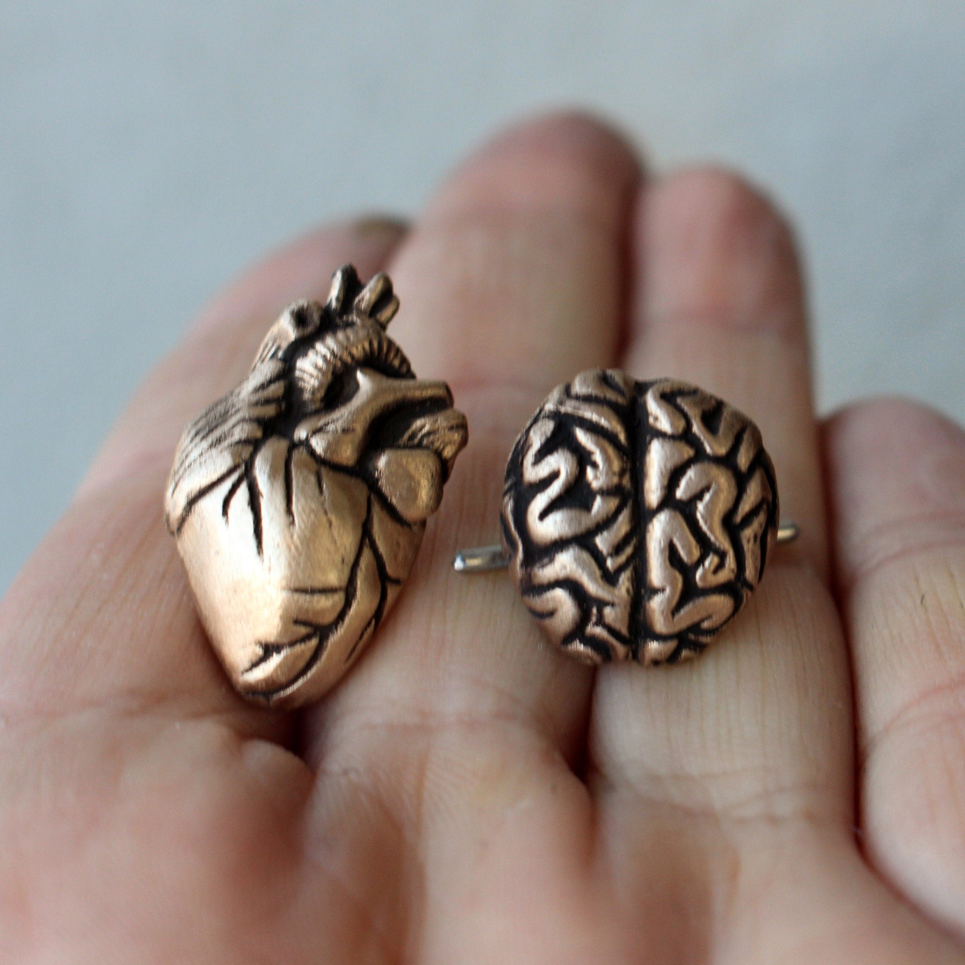 A pair of bronze cufflinks, one shaped like a heart and the other like a brain, held in a person's hand.