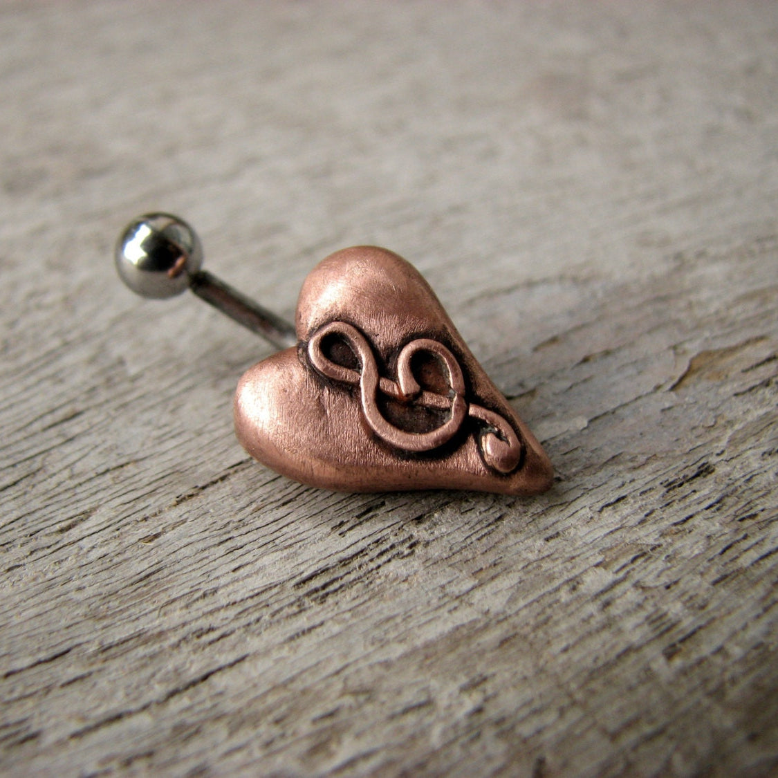 A belly button ring with a puffy heart in pink copper and a g clef design on it, attached to a curved barbell.