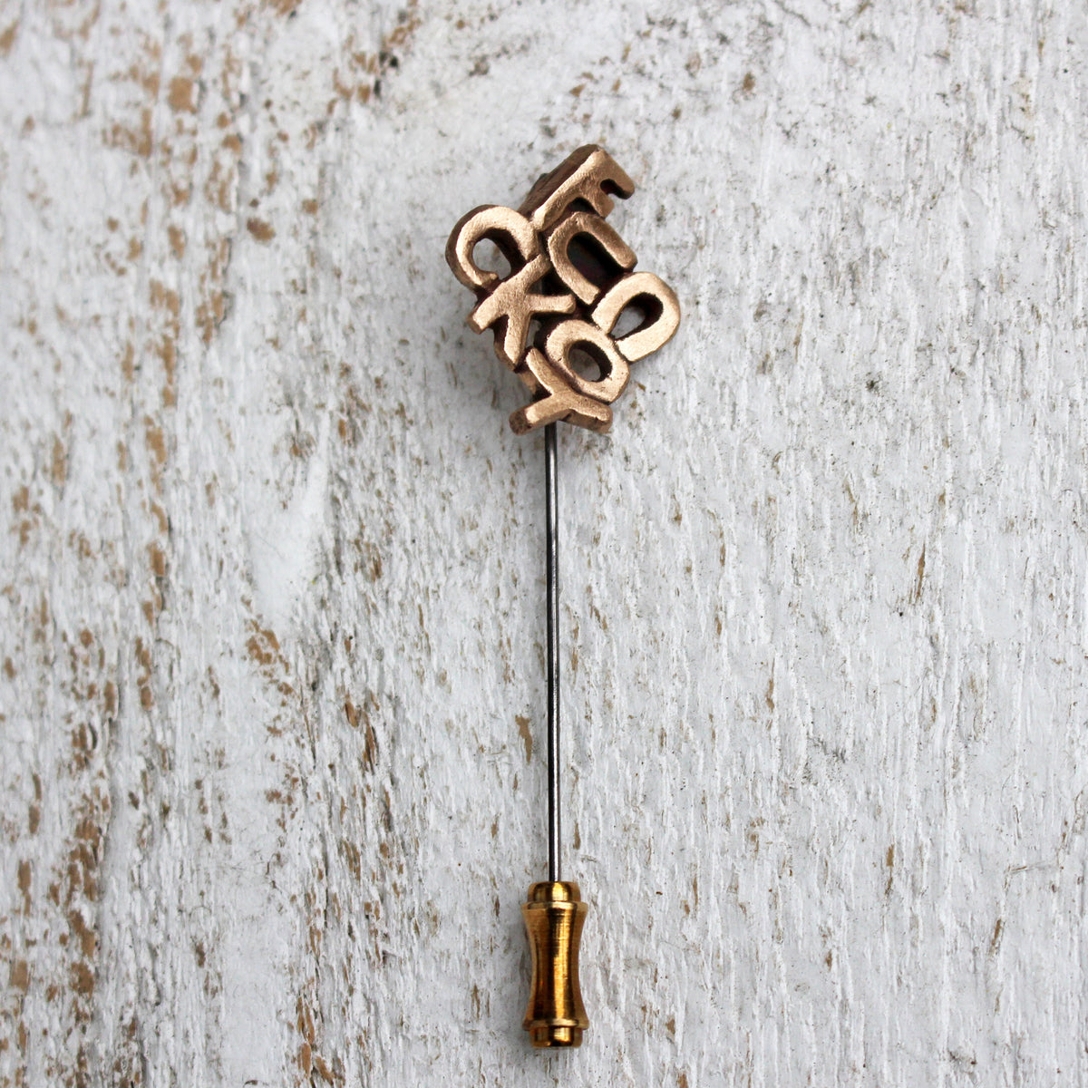 A bronze-colored stick pin with the words 'FUCK YOU' written in stylized lettering protruding from the pin, set against a wooden background.