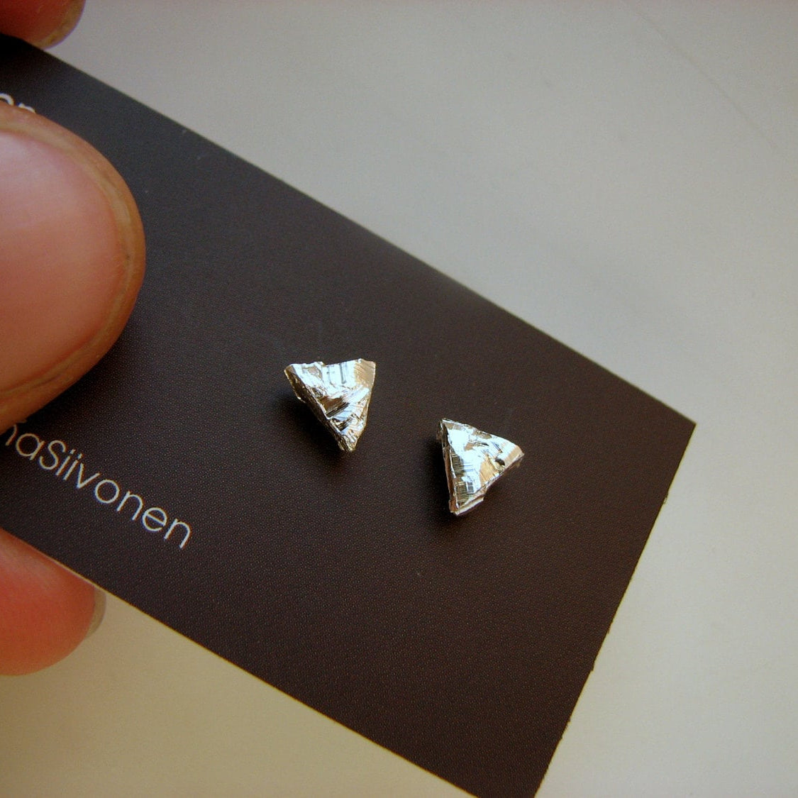 Triangle-shaped silver and titanum earrings on a black card held by a hand