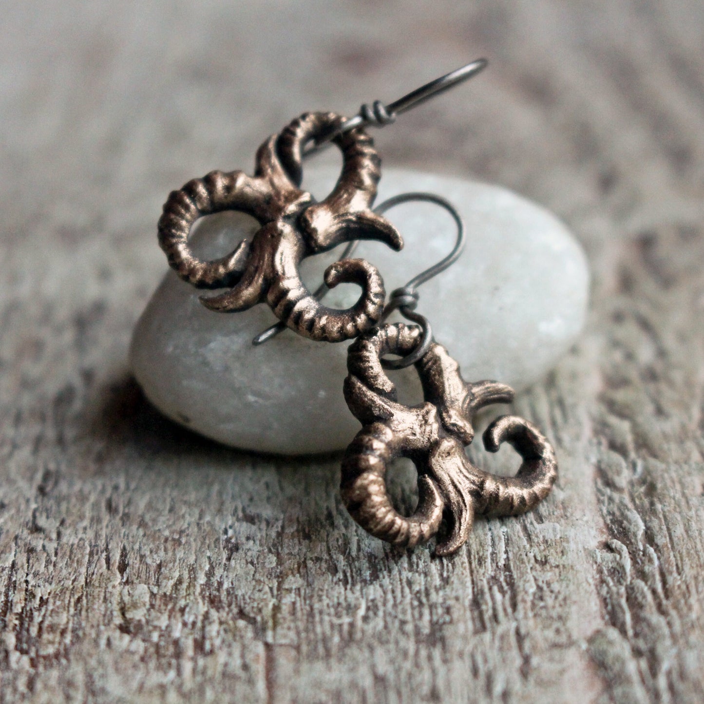 A pair of handmade triskelion earrings made of bronze with hypoallergenic titanium ear wires, displayed on a wooden surface next to a white stone.