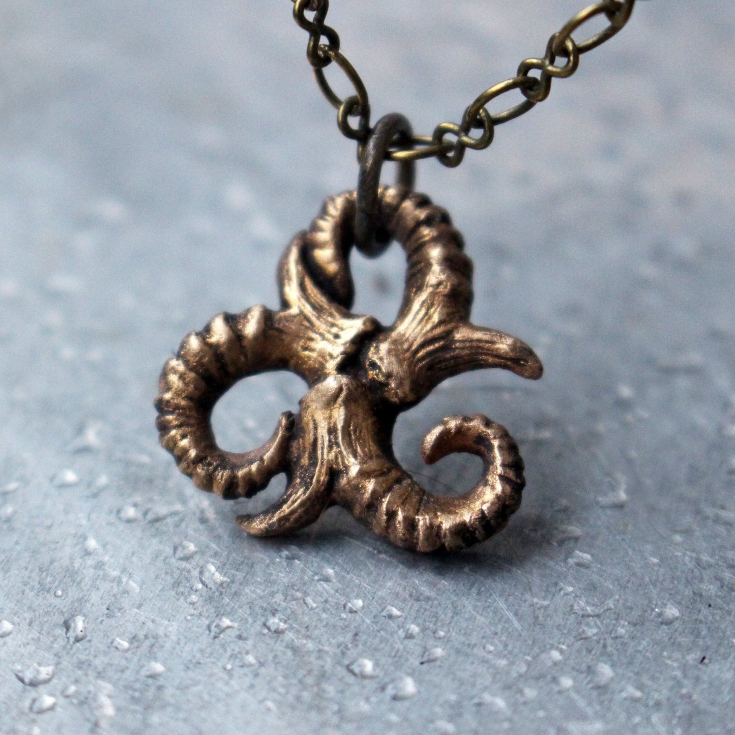 A bronze triskelion necklace with an oxidized and polished finish, displayed on a chain against a textured background.