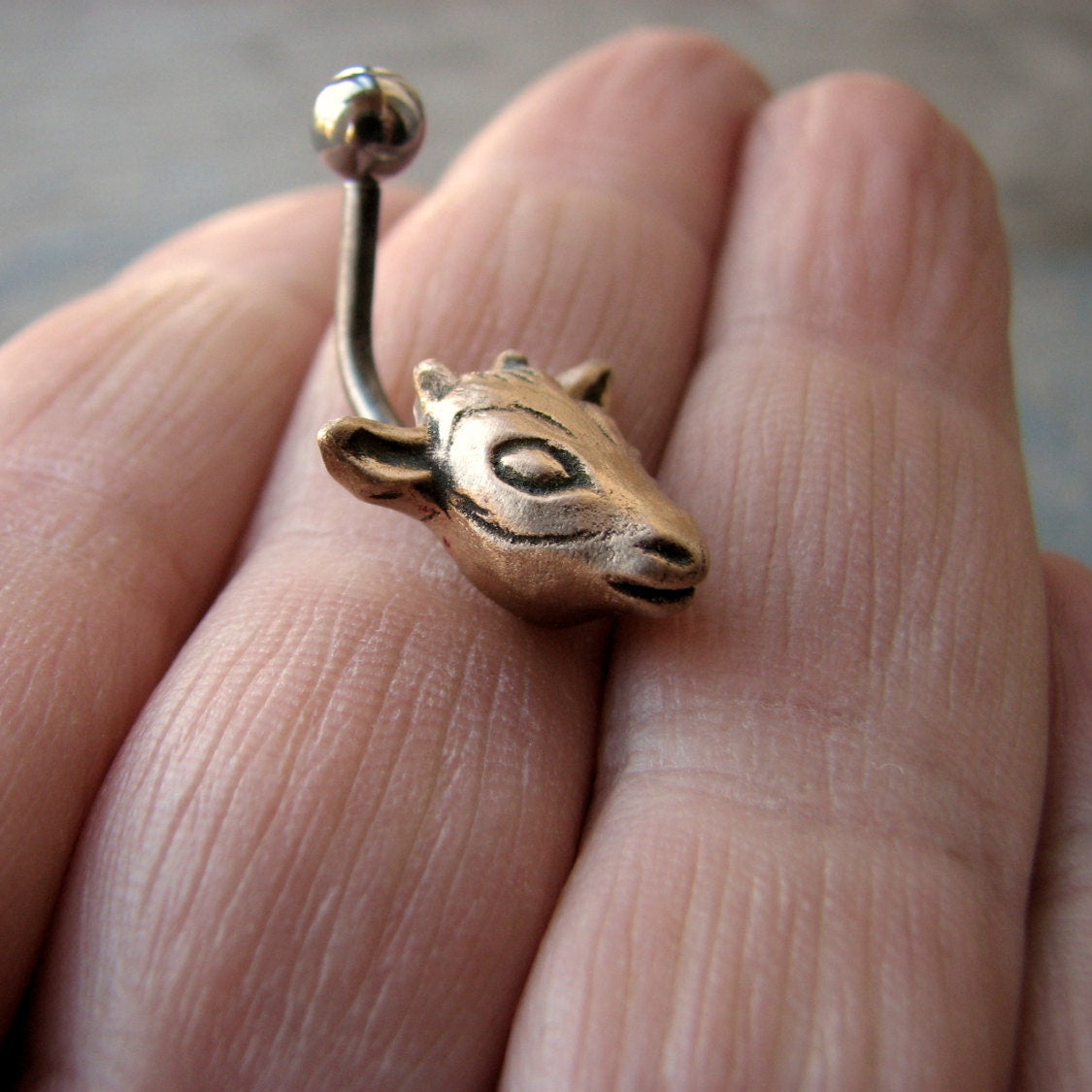 A naval piercing ring with a bronze goat head attached to a barbell, held between fingers.