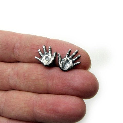 A pair of silver earrings shaped like miniature hands, held between fingers for scale.