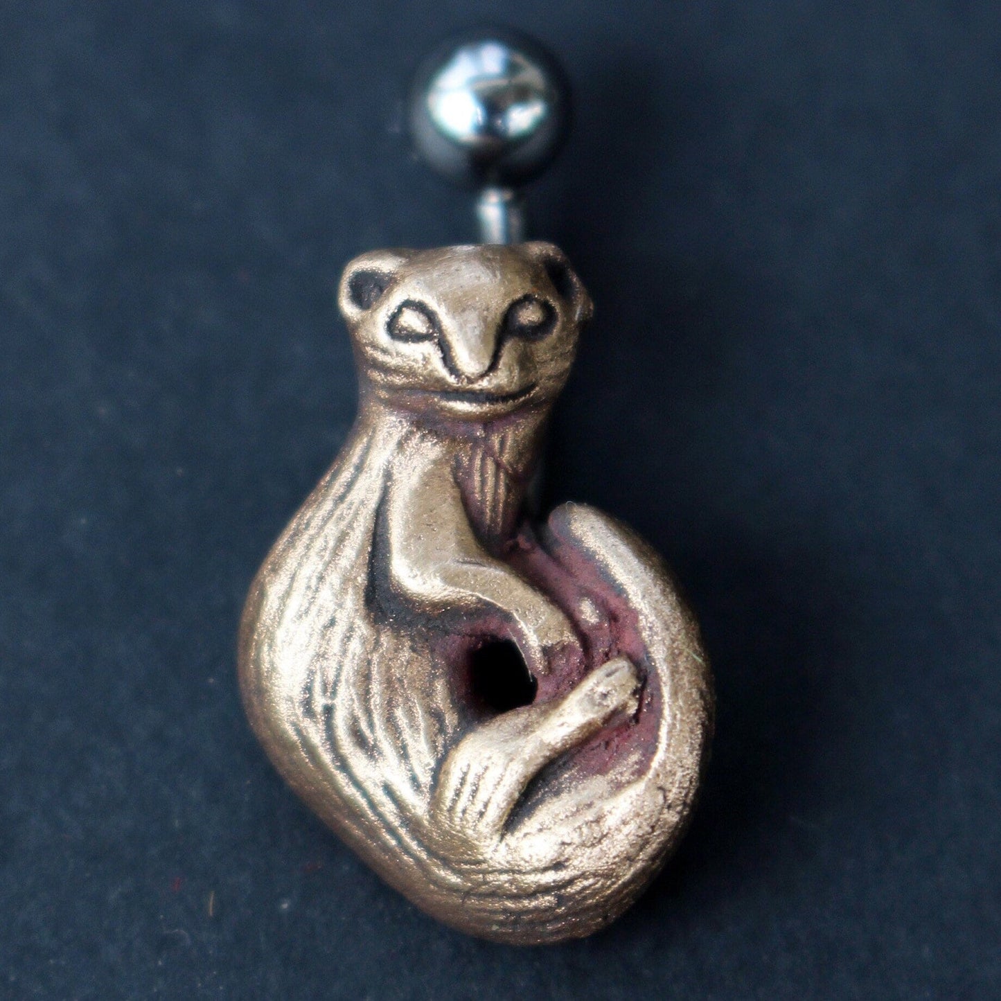 belly button ring with a bronze ferret sculpture on the end, attached to a curved barbell which is presumably made of titanium or surgical steel, set against a dark background.