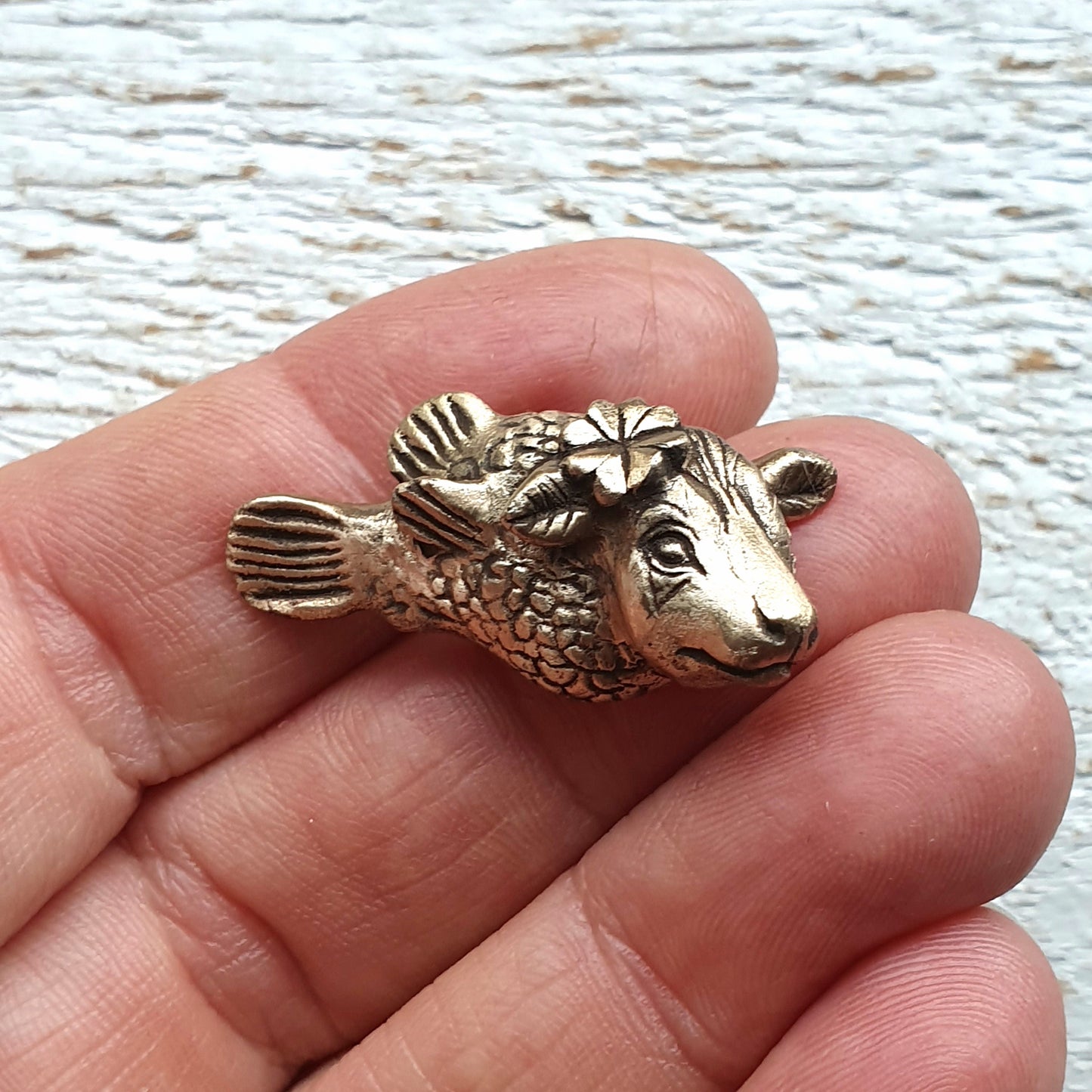 kofisk skulpterat i brons, smycke med en ko med fiskkropp med en fyrklöver bakom ena örat, hålld i en hand